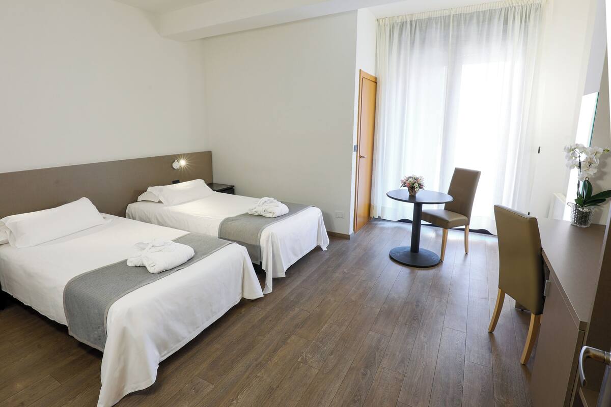 Hotellihuone, jossa on kaksi sänkyä, pieni pyöreä pöytä ja tuoli sekä ikkuna, jossa on verhot.