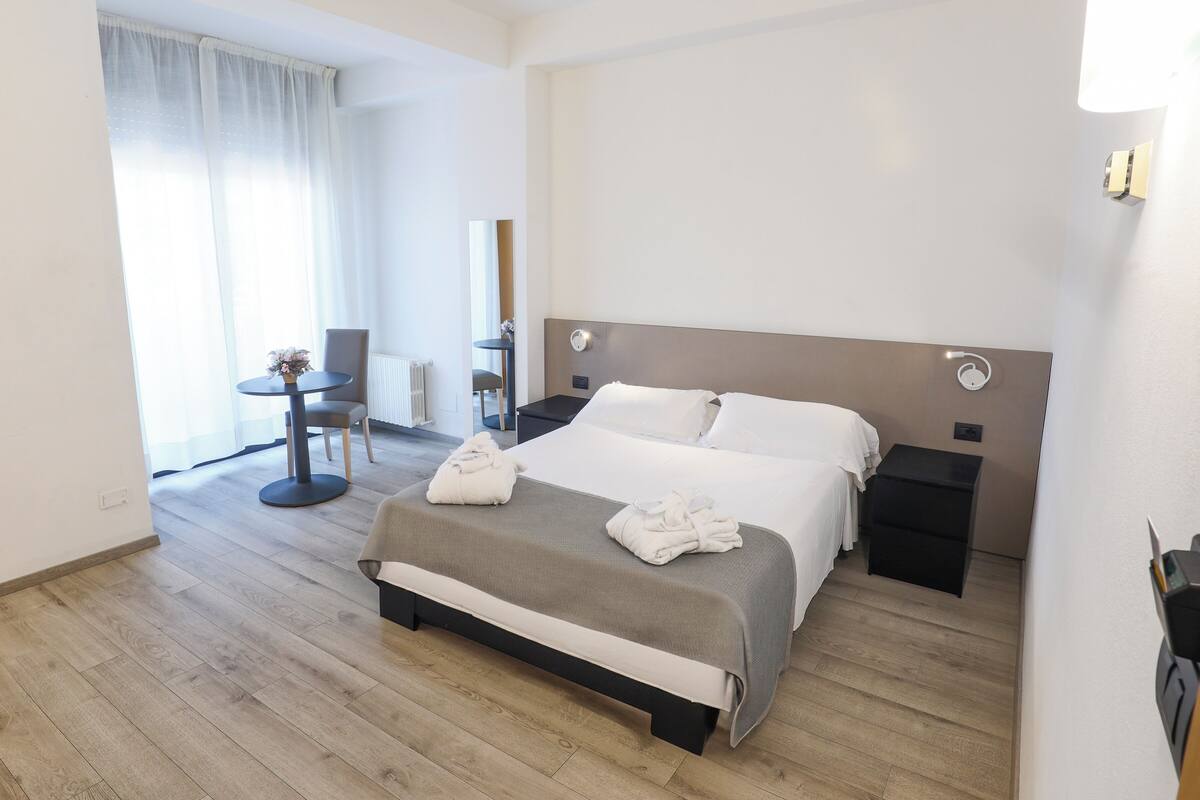 Hotellihuone, jossa on suuri sänky, yöpöydät, pieni pyöreä pöytä ja tuoli, patteri ja ikkuna, jossa on verhot.