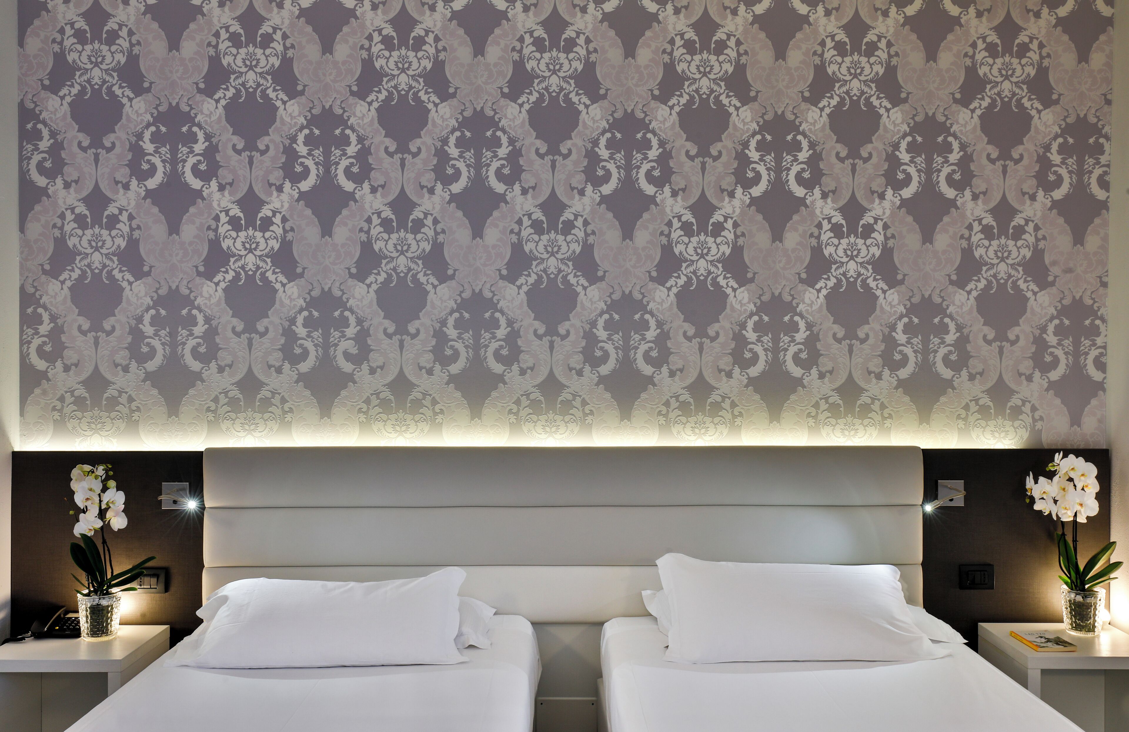 A hotel room with two beds, a patterned wallpaper, and a nightstand with a vase of flowers.