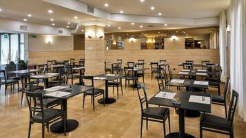 Un'ampia sala ristorante con numerosi tavoli e sedie, una zona cucina sullo sfondo e un interno pulito e moderno.