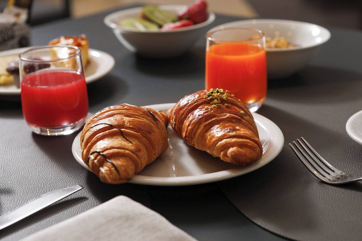 Aamiaispöytä, jossa on croissantteja lautasella, lasi appelsiinimehua ja kulho täynnä hedelmiä.