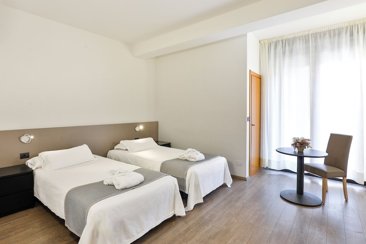 Hotellihuone, jossa on kaksi sänkyä, pieni pöytä ja tuoli sekä ikkuna, jossa on verhot.