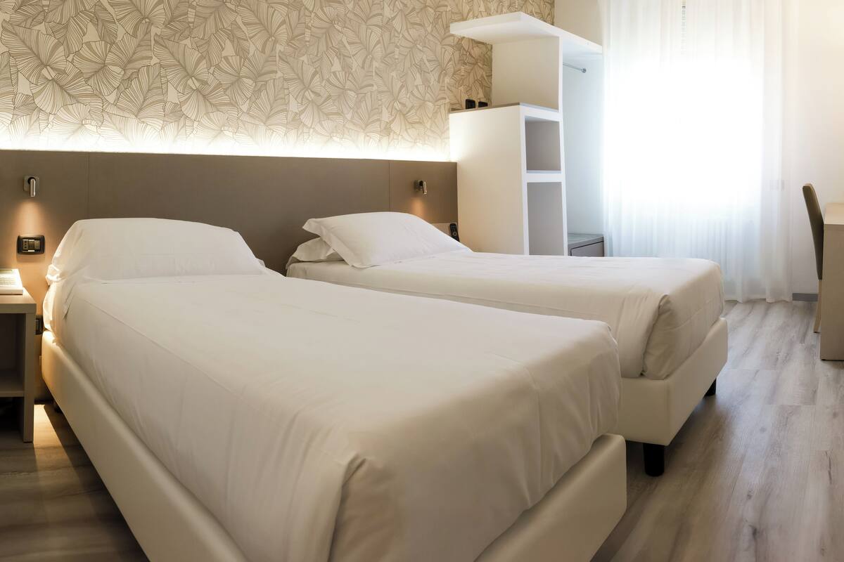 Hotellihuone, jossa on kaksi erillistä sänkyä, työpöytä, tuoli, pieni roskakori ja kuviollinen tapetti.