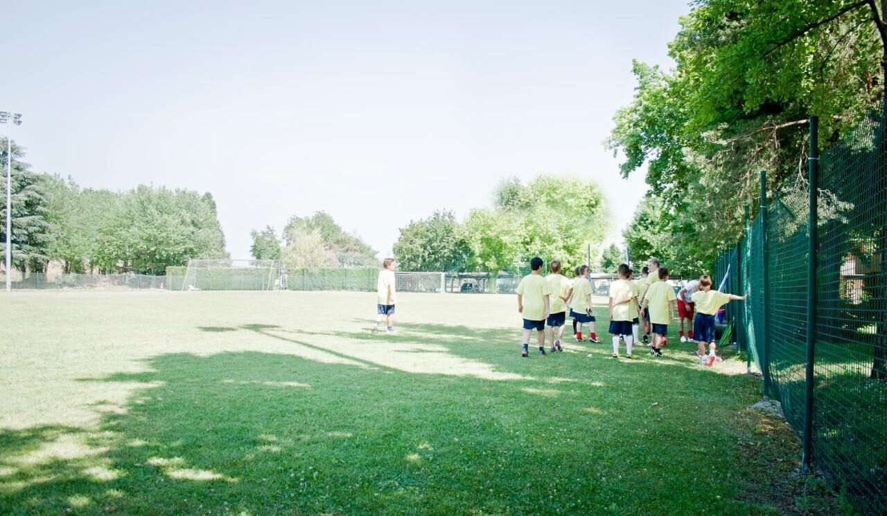 Eine Gruppe von Kindern spielt Fußball auf einer Wiese, im Hintergrund ein Zaun und Bäume.
