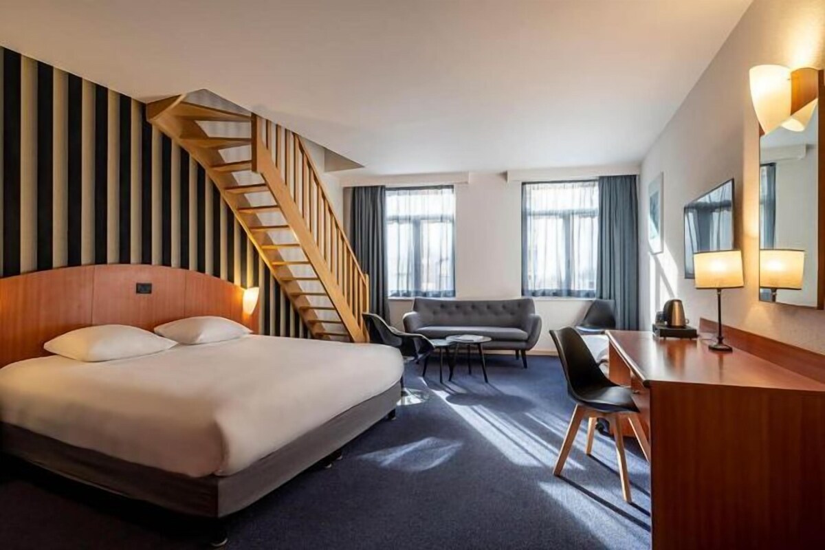 A hotel room with a bed, a desk, a chair, a sofa, a staircase, and a large window.