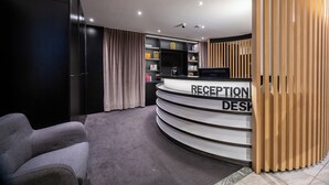 A reception desk with a modern design, a seating area with a grey sofa, and a patterned carpet. - Aris Grand-Place Hotel (Brussels)