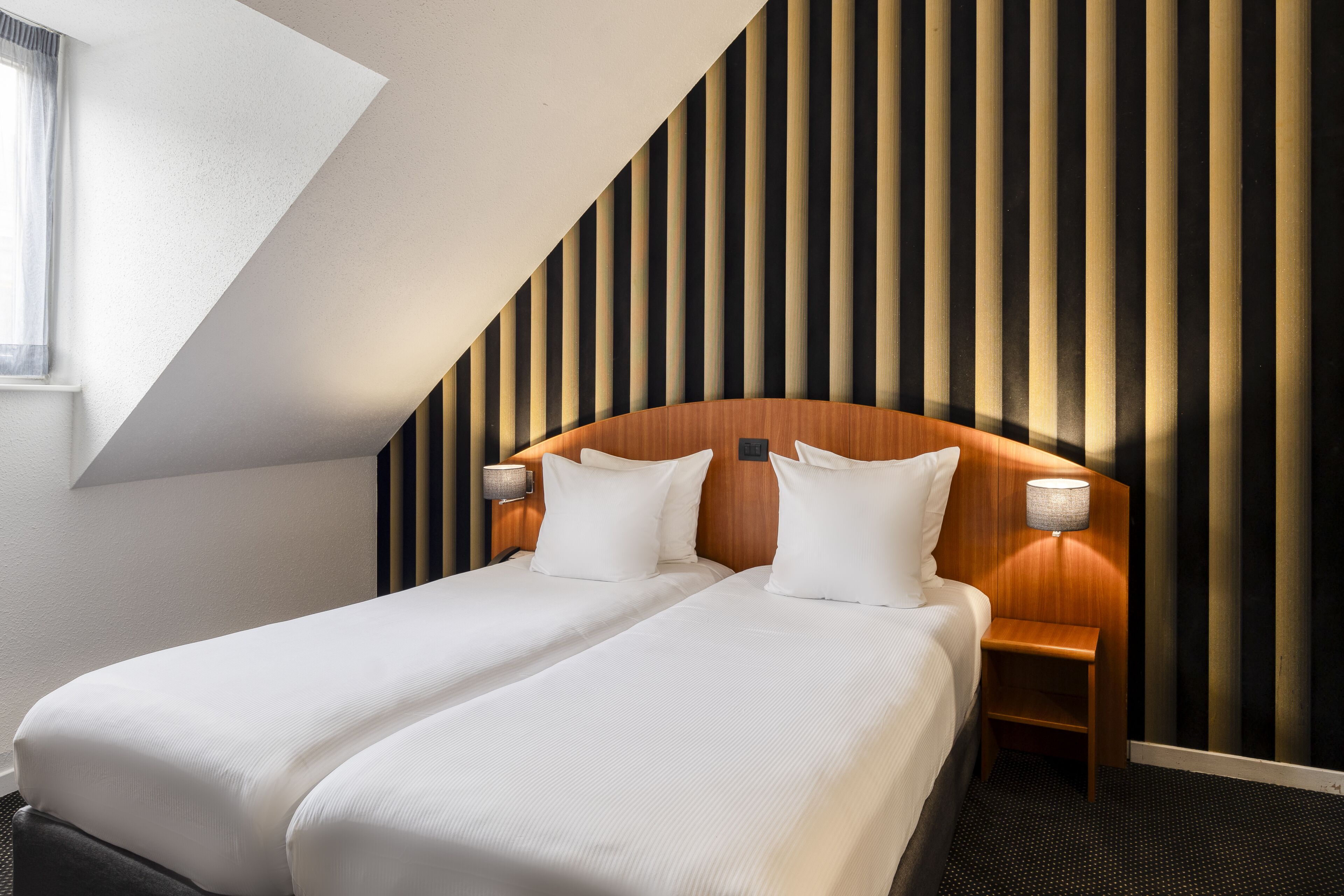Een moderne hotelkamer met een schuin dak, een houten hoofdeinde, twee bedden met witte lakens, een nachtkastje met een lamp, en een gestreept wandontwerp.