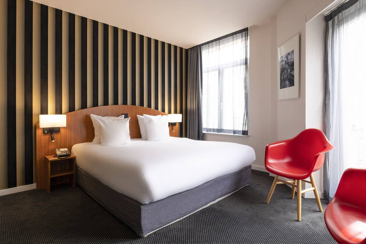 A hotel room with a large bed, a wooden headboard, a nightstand with a lamp, a red chair, and a window with curtains.
