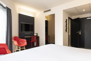 A hotel room with a bed, a TV, a desk, two red chairs, and a black door.