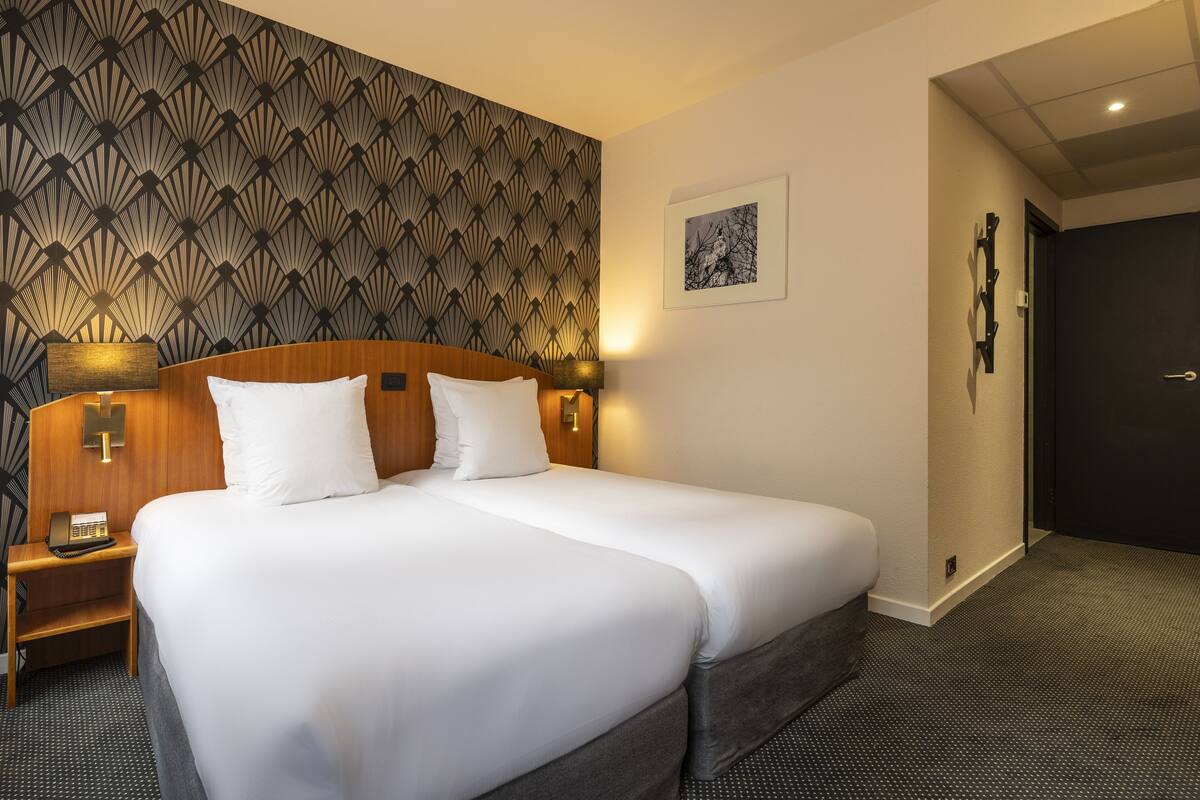 A hotel room with a large bed, a wooden headboard, a bedside table with a lamp, and a patterned wallpaper.