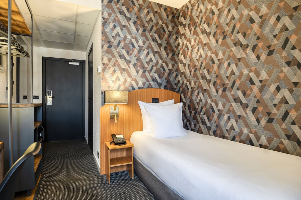 A hotel room with a bed, a desk with a lamp, a telephone, and a door.