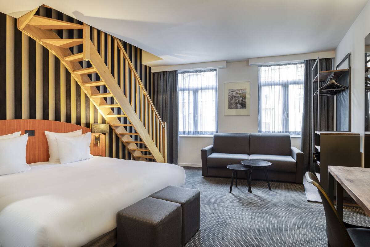 A hotel room with a large bed, a sofa, a small table, a wooden staircase, and a window with curtains.