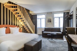 A hotel room with a large bed, a sofa, a small table, a wooden staircase, and a window with curtains.