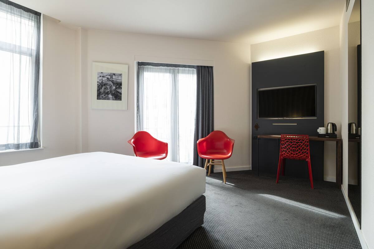 A hotel room with a large bed, two red chairs, a desk with a flat-screen TV, and a framed picture on the wall.