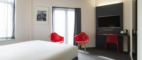 A hotel room with a large bed, two red chairs, a desk with a flat-screen TV, and a framed picture on the wall.