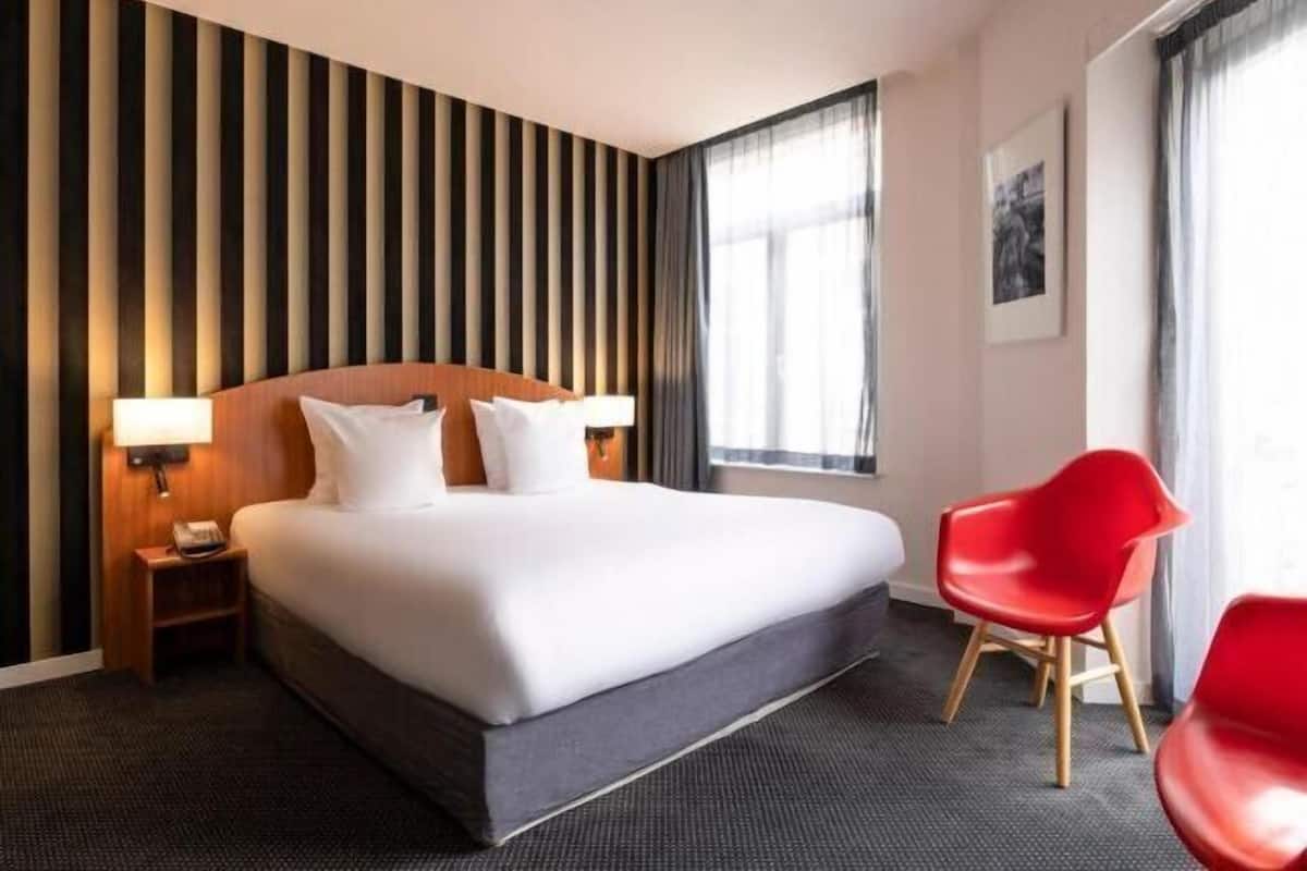A hotel room with a large bed, a nightstand, a red chair, and a striped wall.