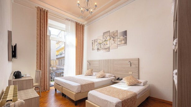 A hotel room with two beds, a desk with a kettle, a chair, and a view of a building outside. - Lilium Hotel (Florence)