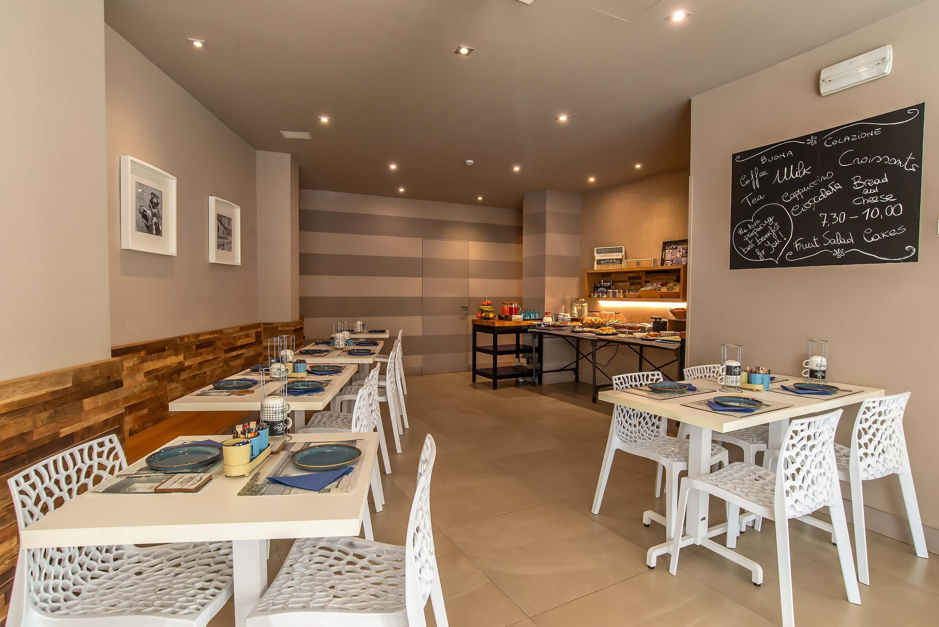 A restaurant with a long dining area, white chairs, and tables set with plates and cutlery. A chalkboard menu is on the wall listing food and drink options.