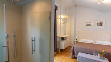 Camera d'albergo con doccia, un letto con comodino, una lampada da comodino, un attaccapanni e un piccolo tavolino con una bottiglia e dei bicchieri.