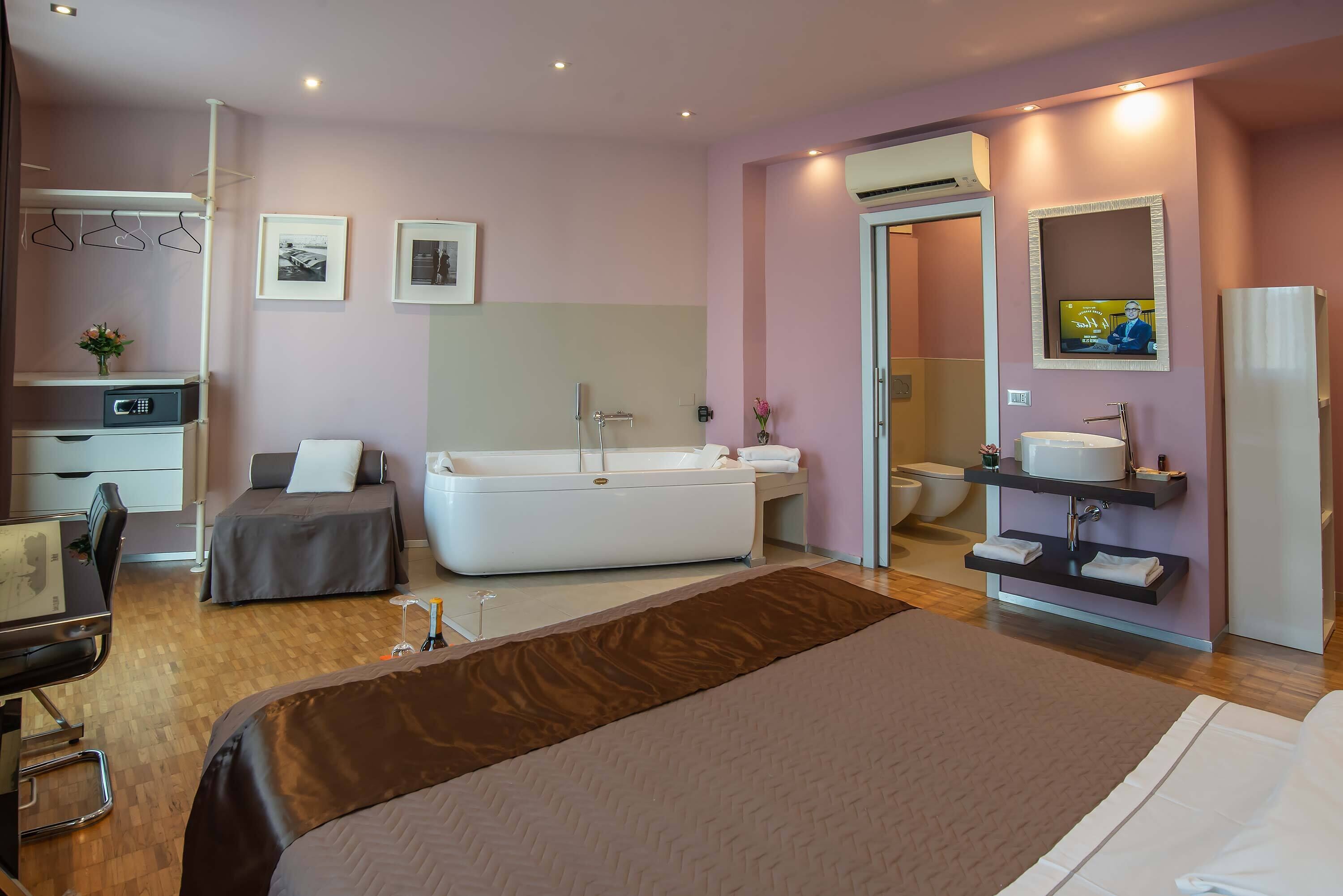 A modern hotel room with a large bed, a sitting area with a desk and chair, a bathtub, a vanity with a mirror, and a toilet.