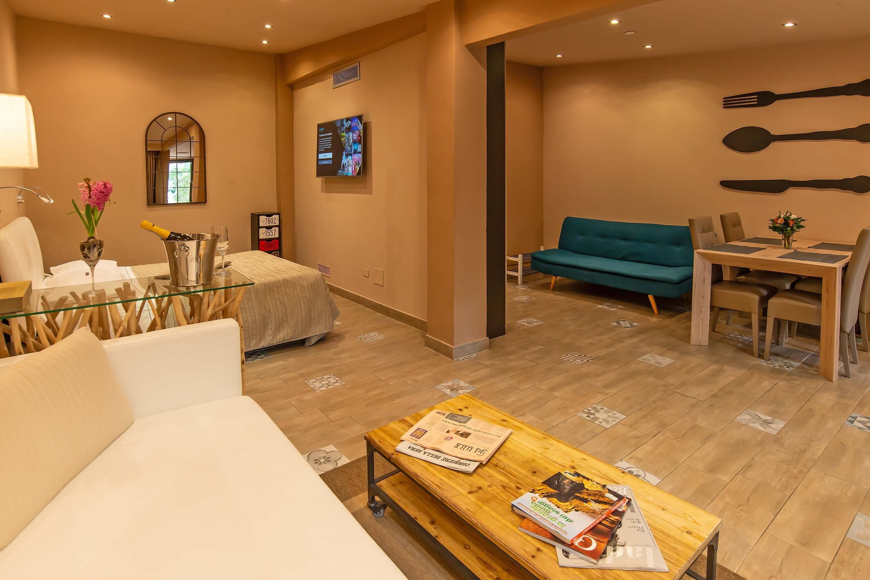 A modern hotel room with a glass coffee table, a sofa, a dining area with a table and chairs, and a TV mounted on the wall.
