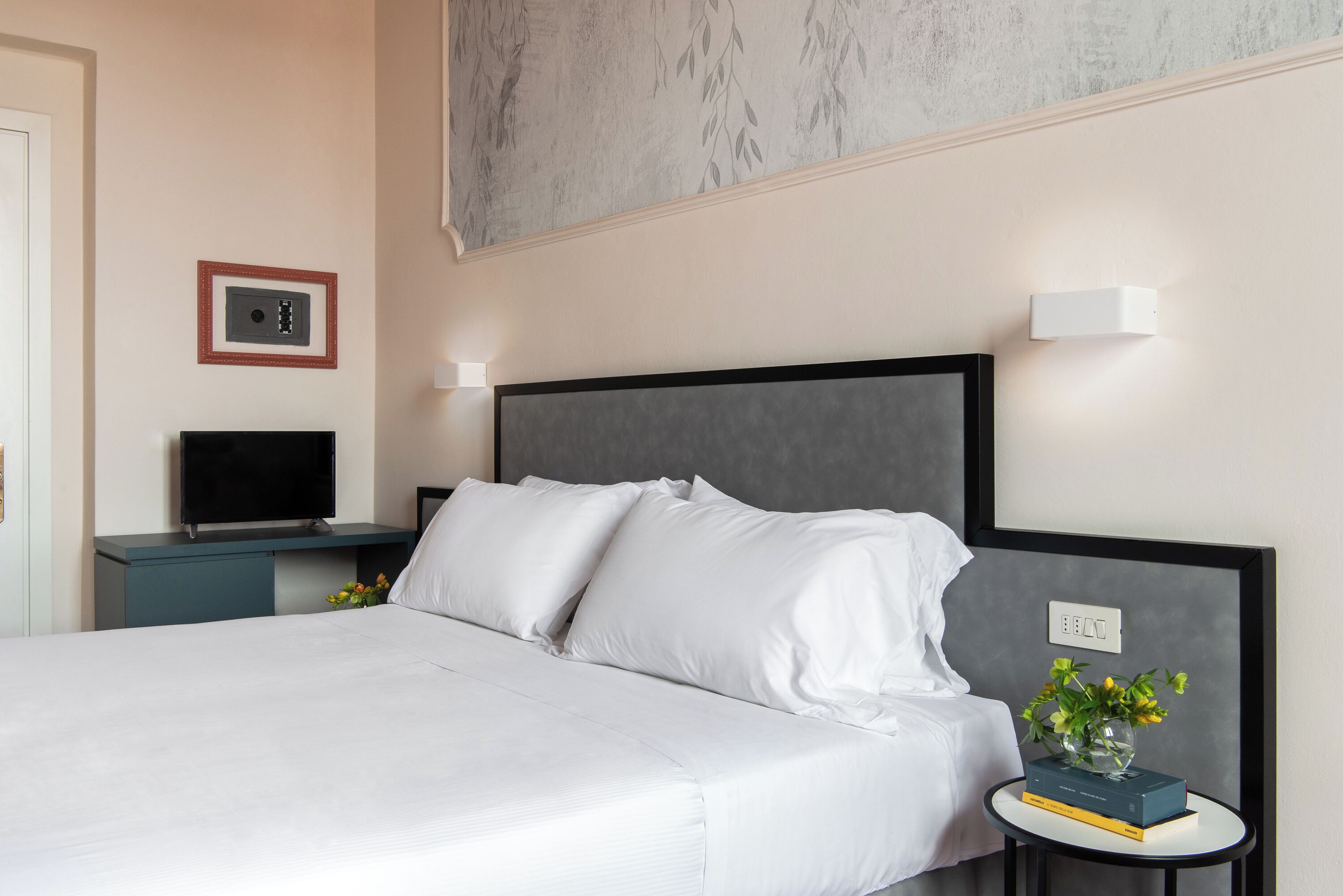 A neatly made bed with white linens, a small bedside table with a book and a vase of flowers, and a wall-mounted television.