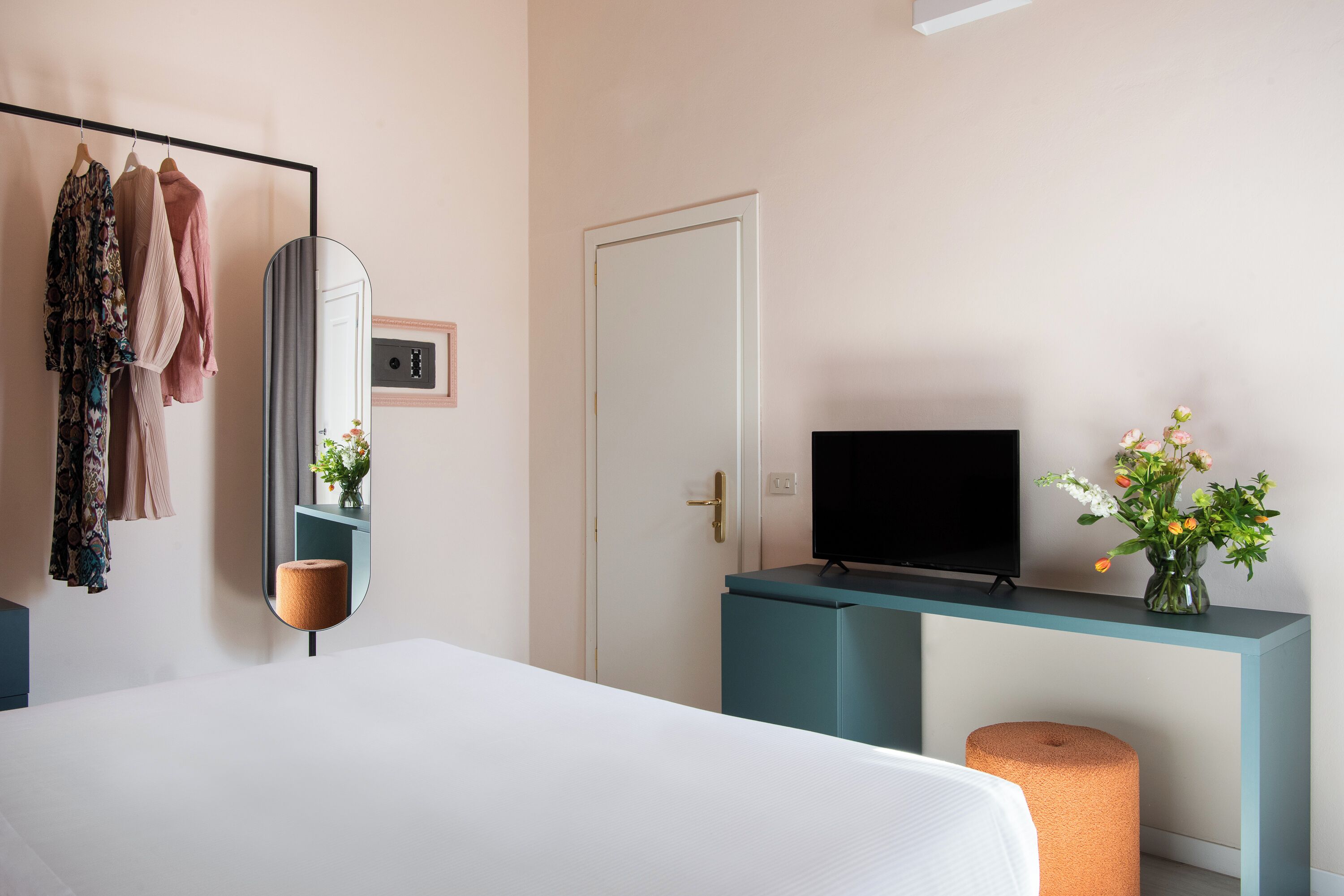 A hotel room with a bed, a TV on a stand, a mirror, a wardrobe, and a small table with a vase of flowers.