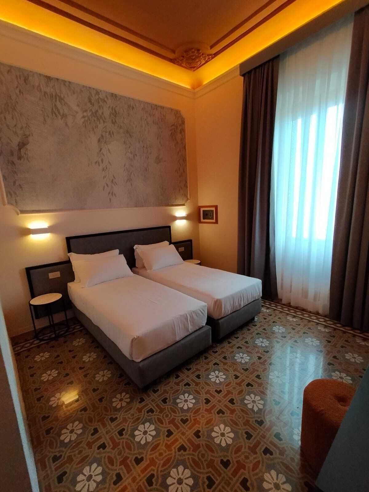 A hotel room with two beds, a patterned floor, a large window with curtains, and a decorative ceiling.