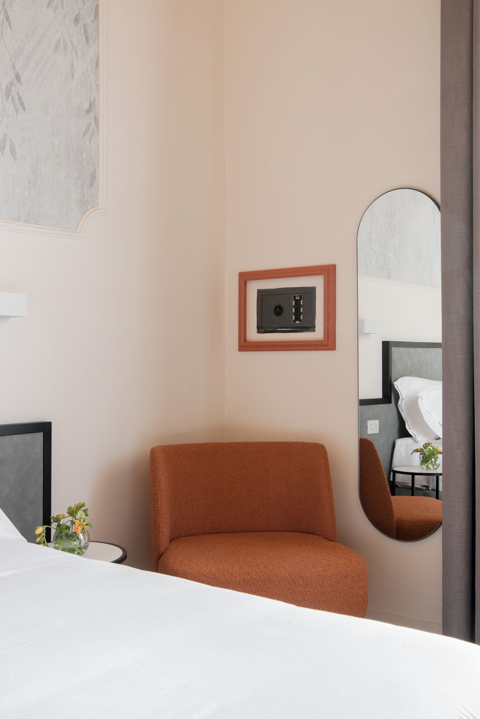 Hotelkamer met een bed, een bruine fauteuil, een ronde spiegel, een wandthermostaat en een lijst met een afbeelding.
