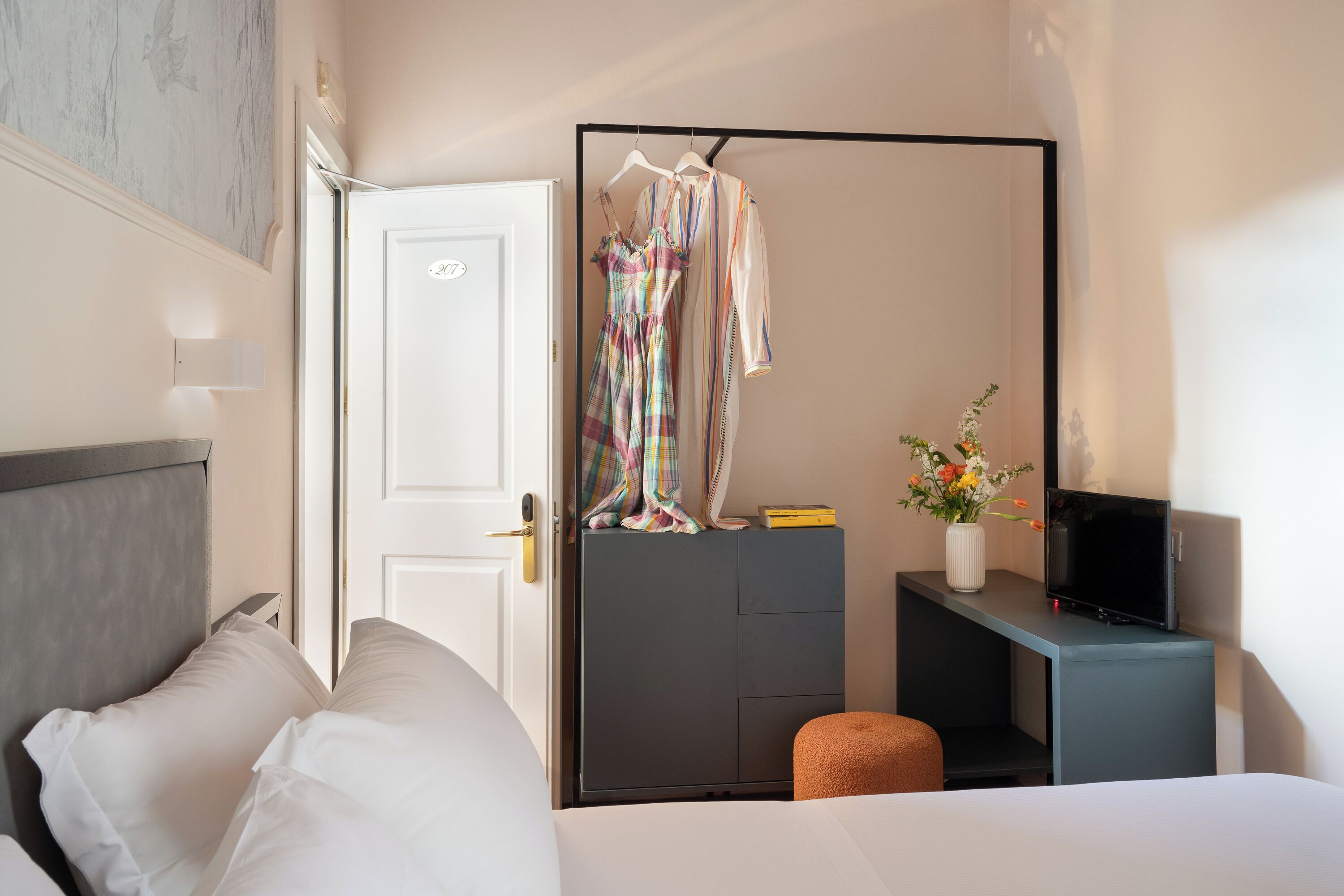 Une chambre d’hôtel comprenant un lit, une armoire, un bureau avec une télévision et une petite table sur laquelle est posé un vase de fleurs.