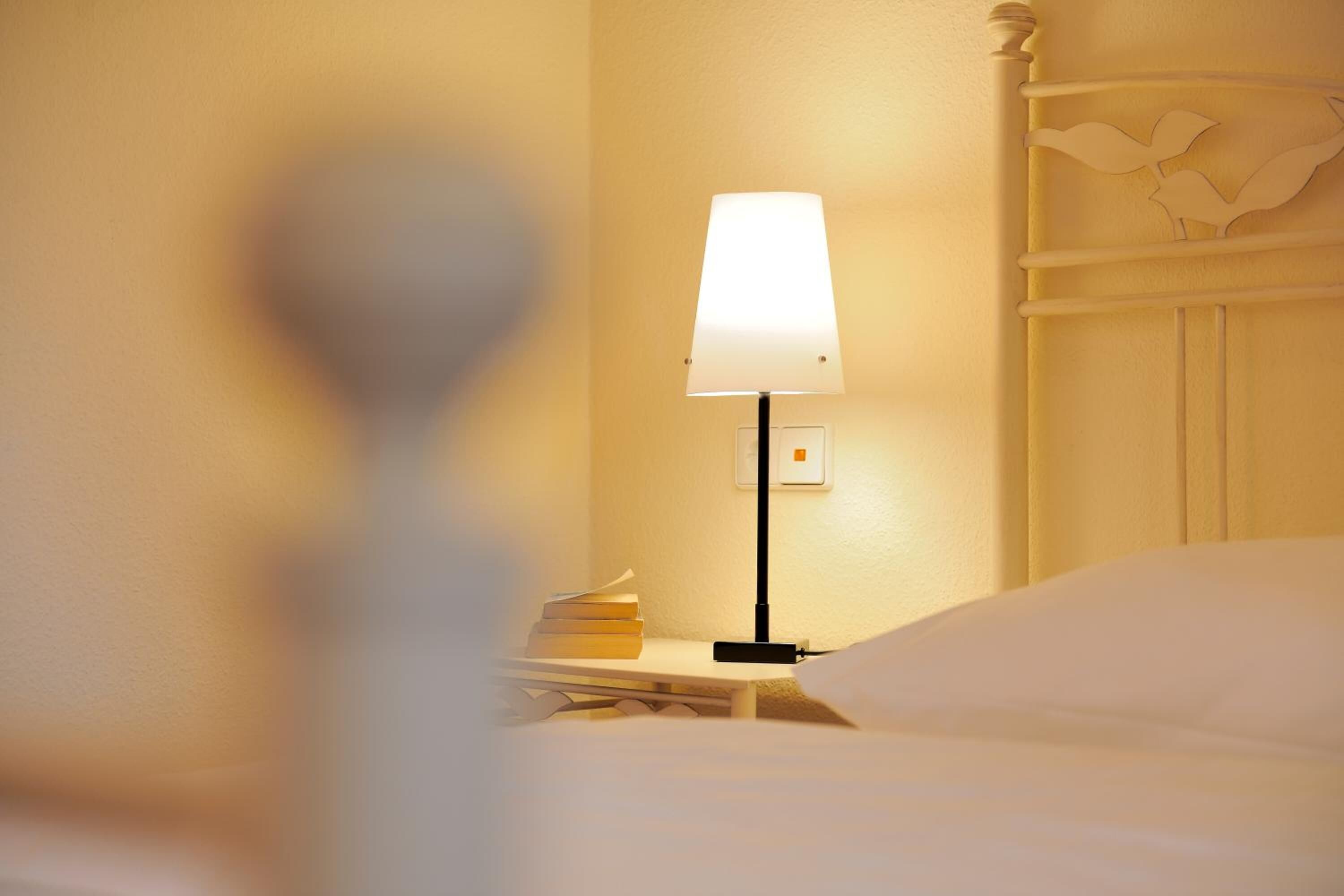 Una cama bien tendida con cabecera blanca, una mesita de noche con lámpara y una pila de libros.