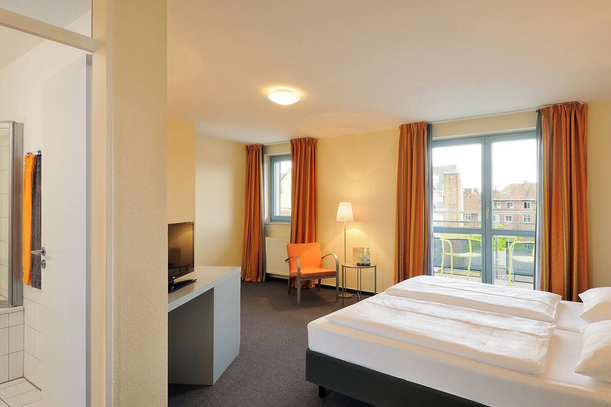 A hotel room with a bed, a desk with a TV, a chair, a window with curtains, and a view of buildings outside.