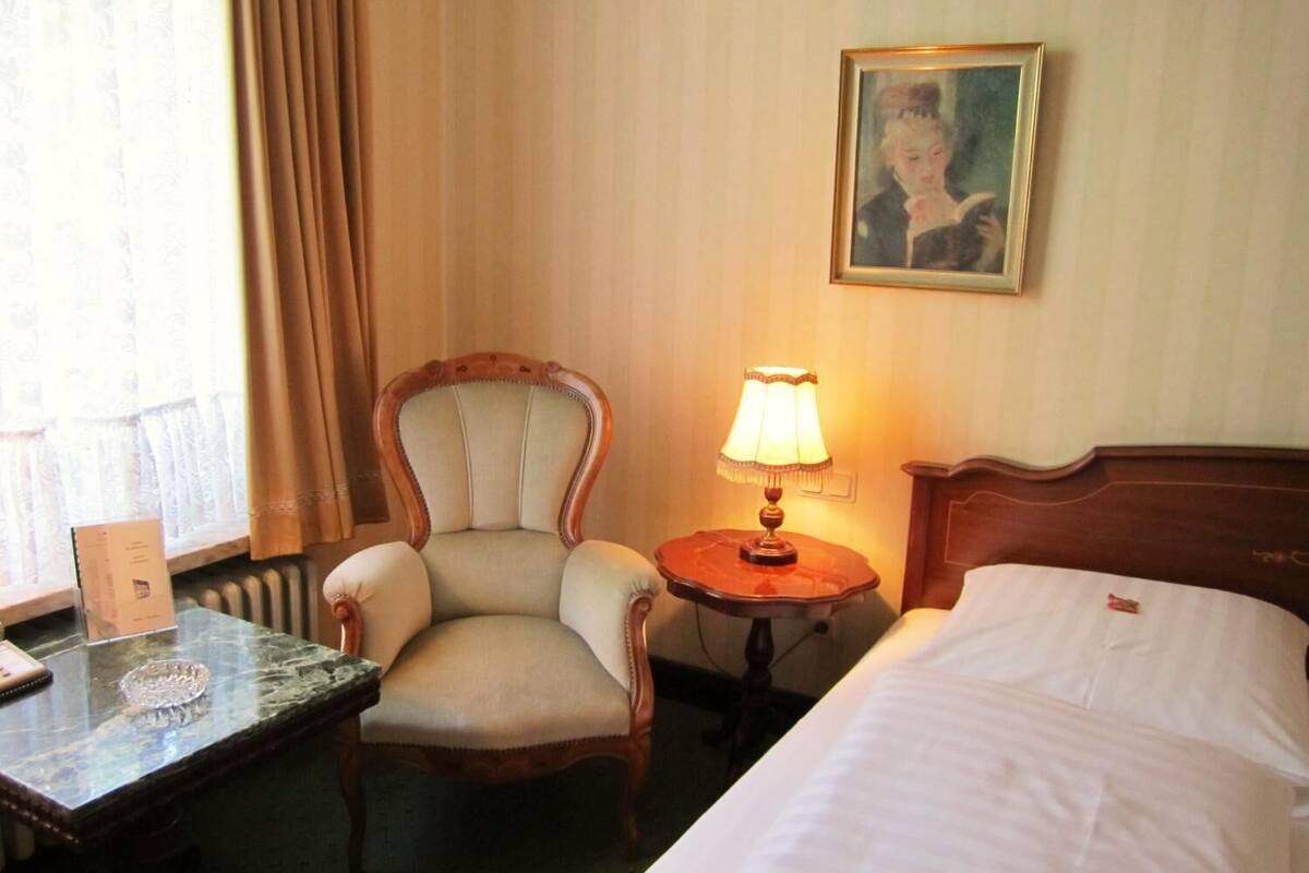 A hotel room with a bed, a chair, a table, a lamp, a painting, and a window with curtains.