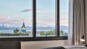 A hotel room with a large window, a bed, and a view of a church and mountains. - Fosshotel Husavik (Husavik)
