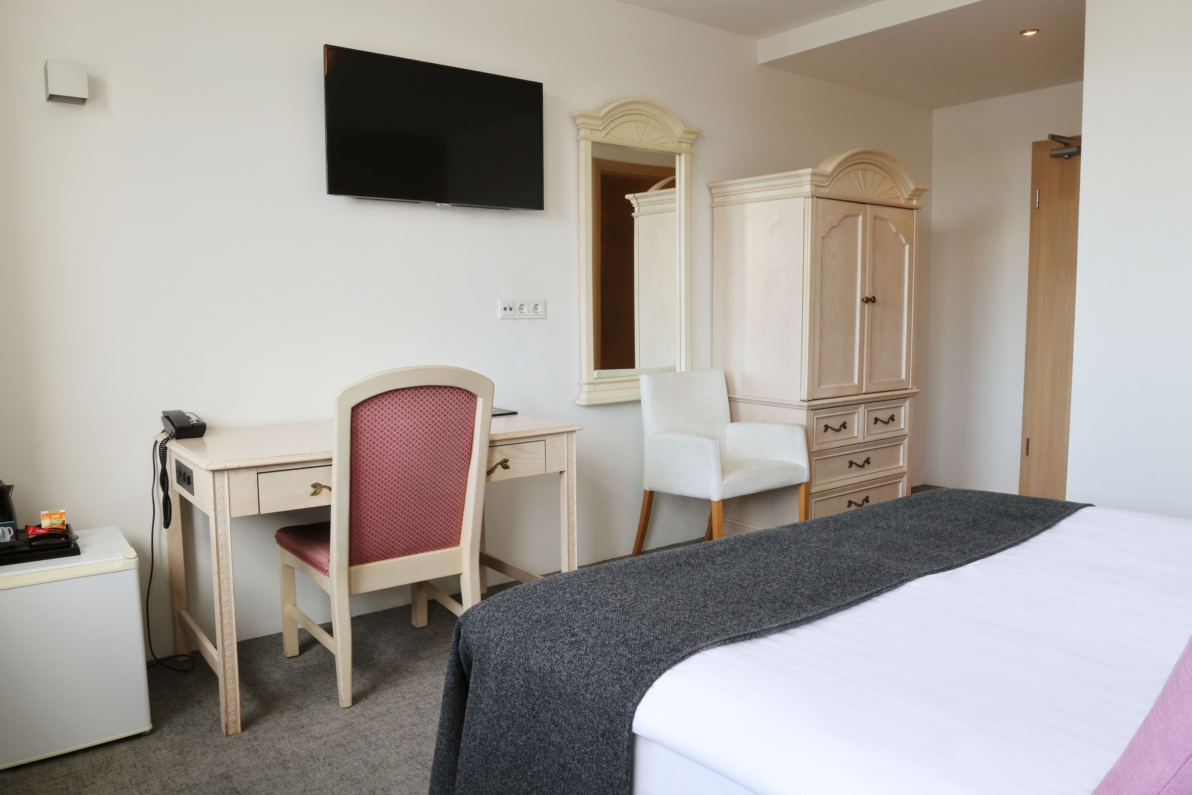 A hotel room with a bed, a desk with a chair, a wardrobe, and a television on the wall.