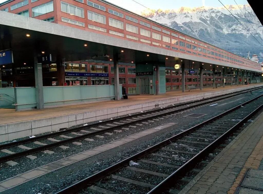 Un androc de tren amb un edifici modern, diverses vies i un paisatge de muntanyes al fons.