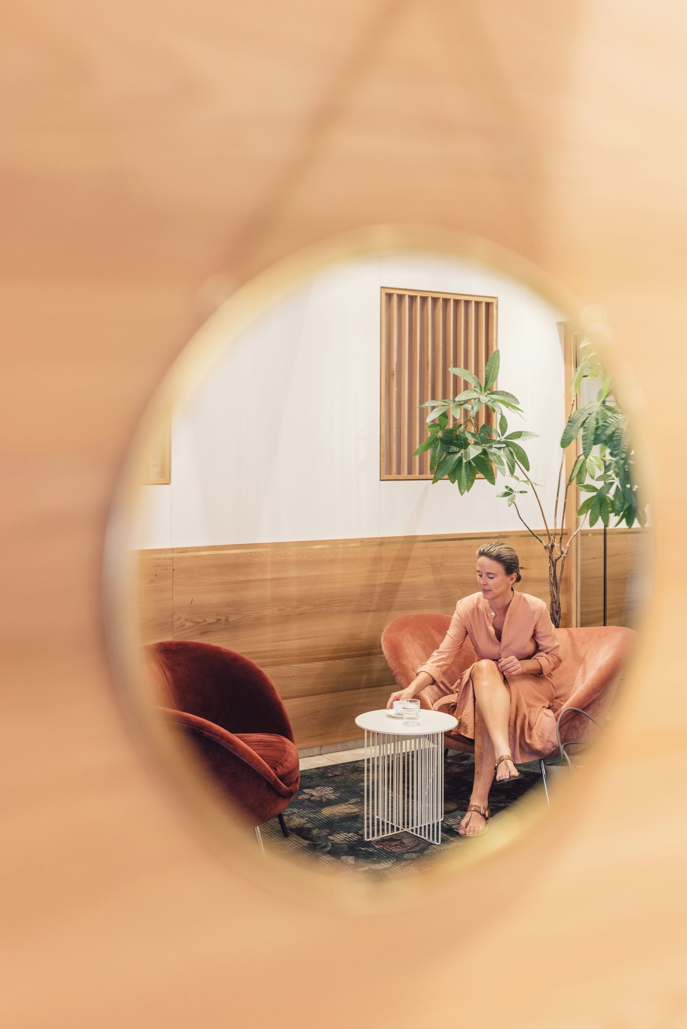 Une personne est assise sur un fauteuil moelleux dans une pièce dont un mur est revêtu de lambris en bois, où se trouvent une petite table ronde et un pot de fleurs.