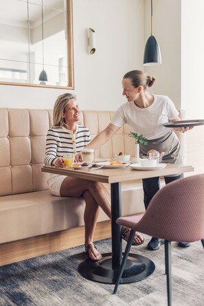 A modern living room with a sofa, a dining table with breakfast items, and a person serving food. - Boutique Hotel Zach (Innsbruck)
