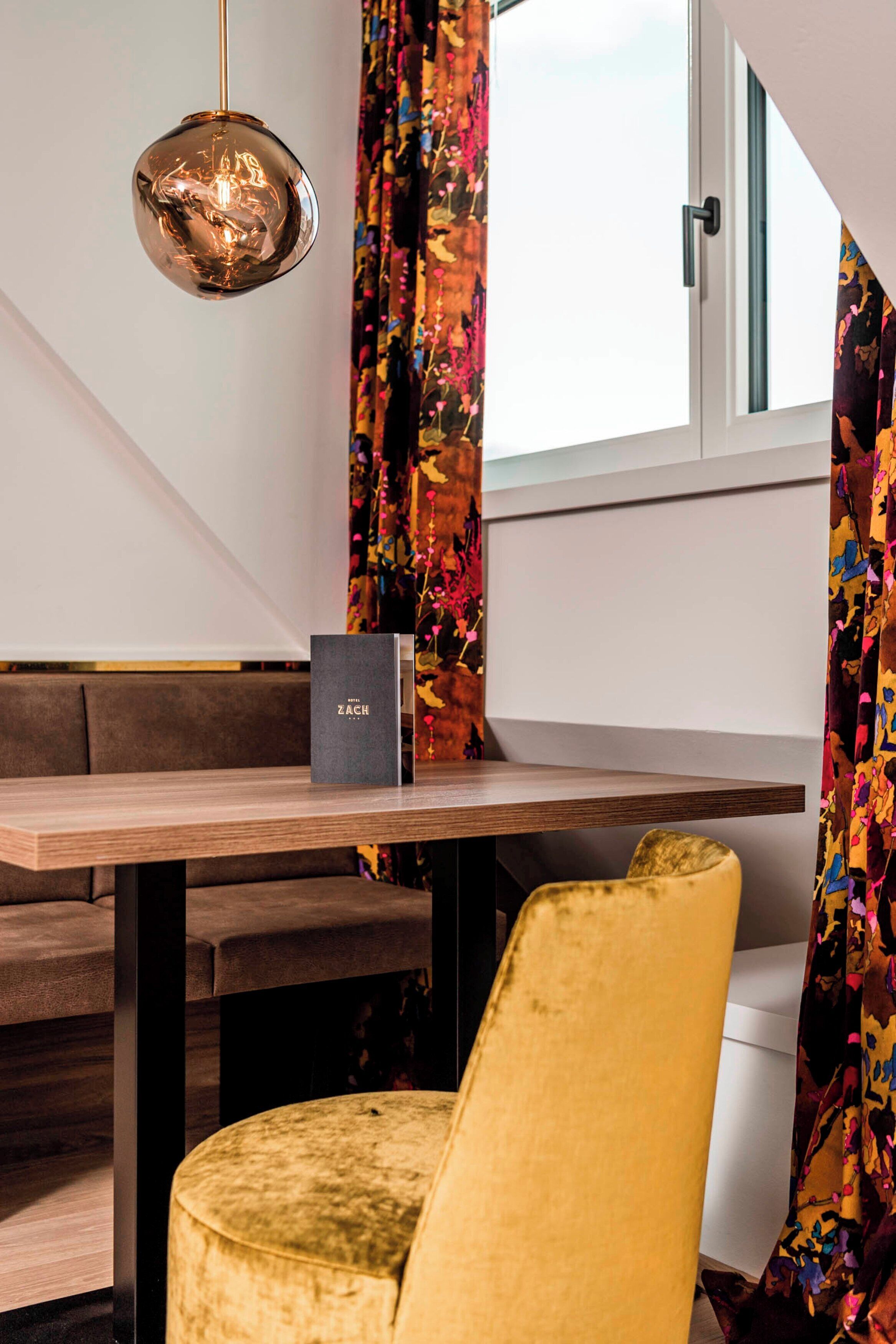 Un intérieur moderne avec une table en bois, un banc, un fauteuil jaune recouvert de tissu, une suspension et une fenêtre avec des rideaux.