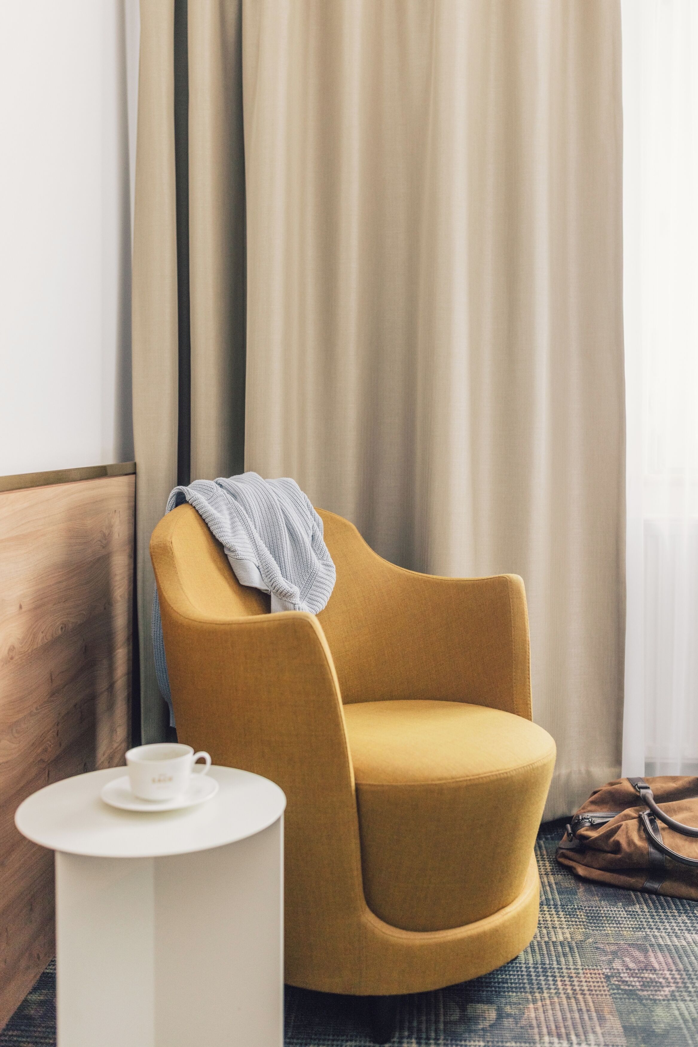 A hotel room with a mustard-colored armchair, a small white side table with a cup, and a brown bag on the floor.