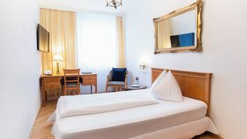 A hotel room with a bed, a desk with a chair, a lamp, a mirror, and a window with curtains.