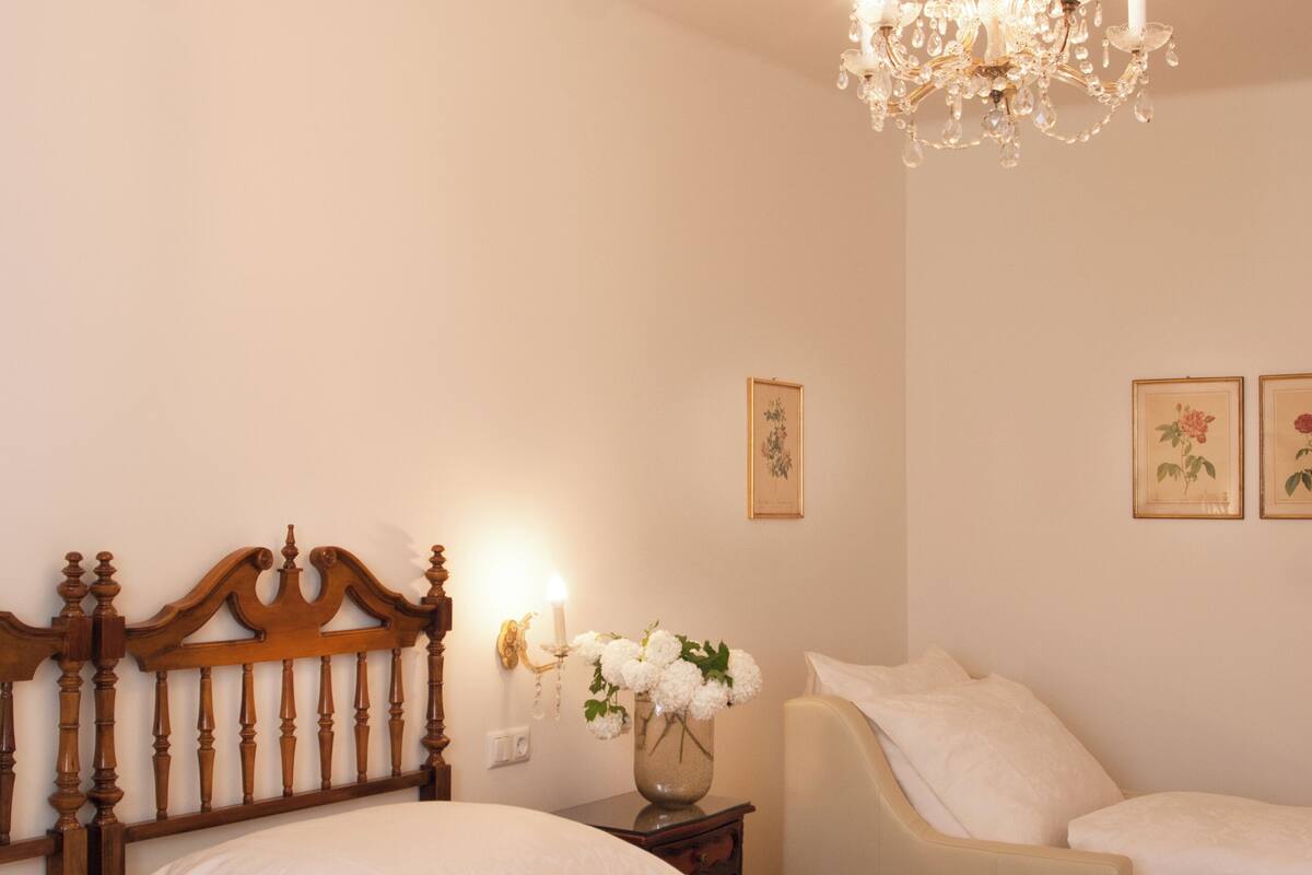 A bedroom with a wooden headboard, a chandelier, a bedside table with a vase of flowers, and two white chairs.