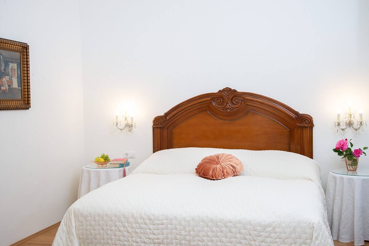 A bedroom with a wooden headboard, a white quilted bedspread, a single pink pillow, a small bedside table with a lamp, and a framed picture on the wall.