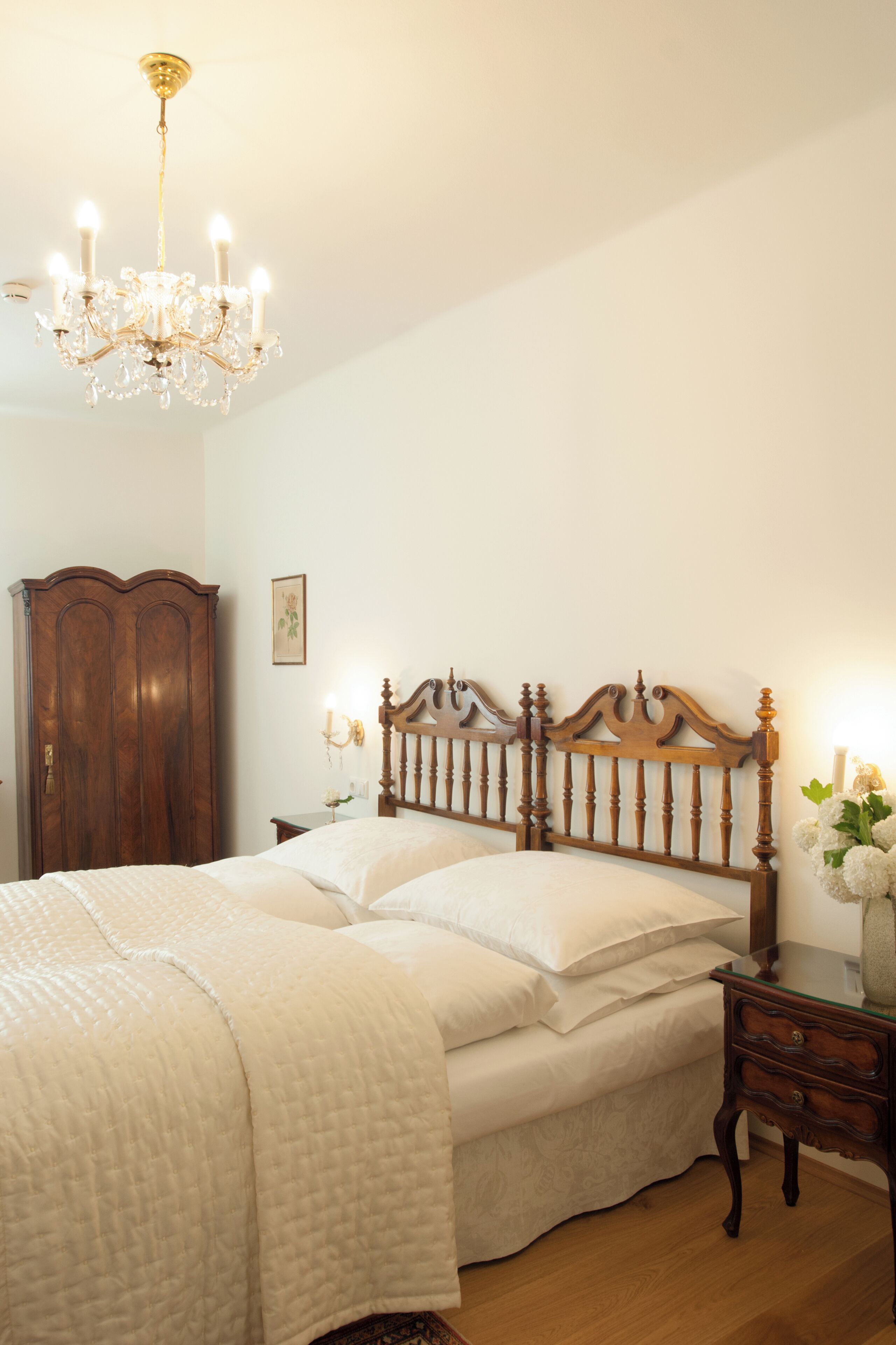 A bedroom with a wooden bed, a chandelier, a wooden wardrobe, and a bedside table with a vase of flowers.