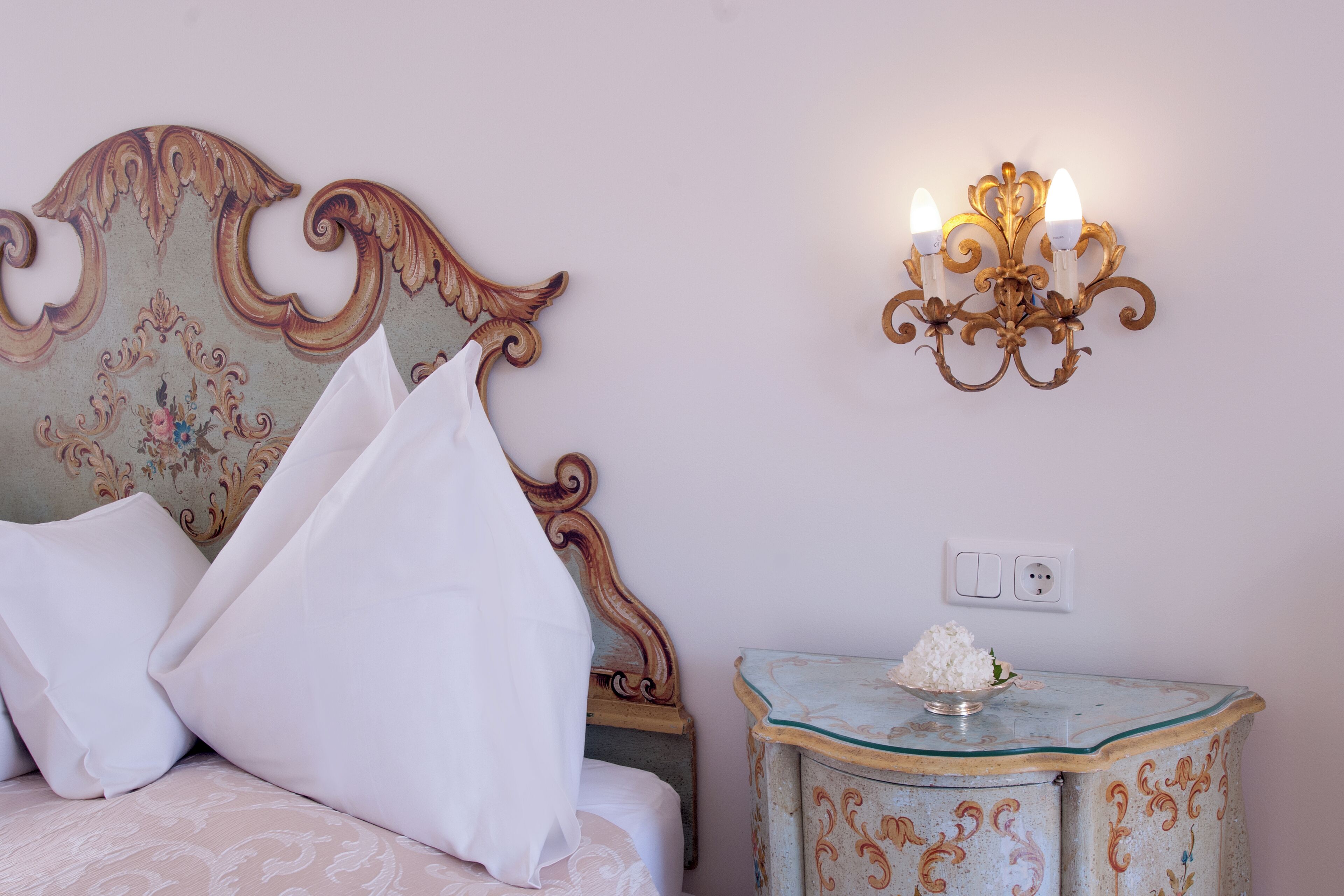 A bedroom with a bed, white pillows, a decorative headboard, a bedside table with a bowl of flowers, and a wall-mounted light fixture.