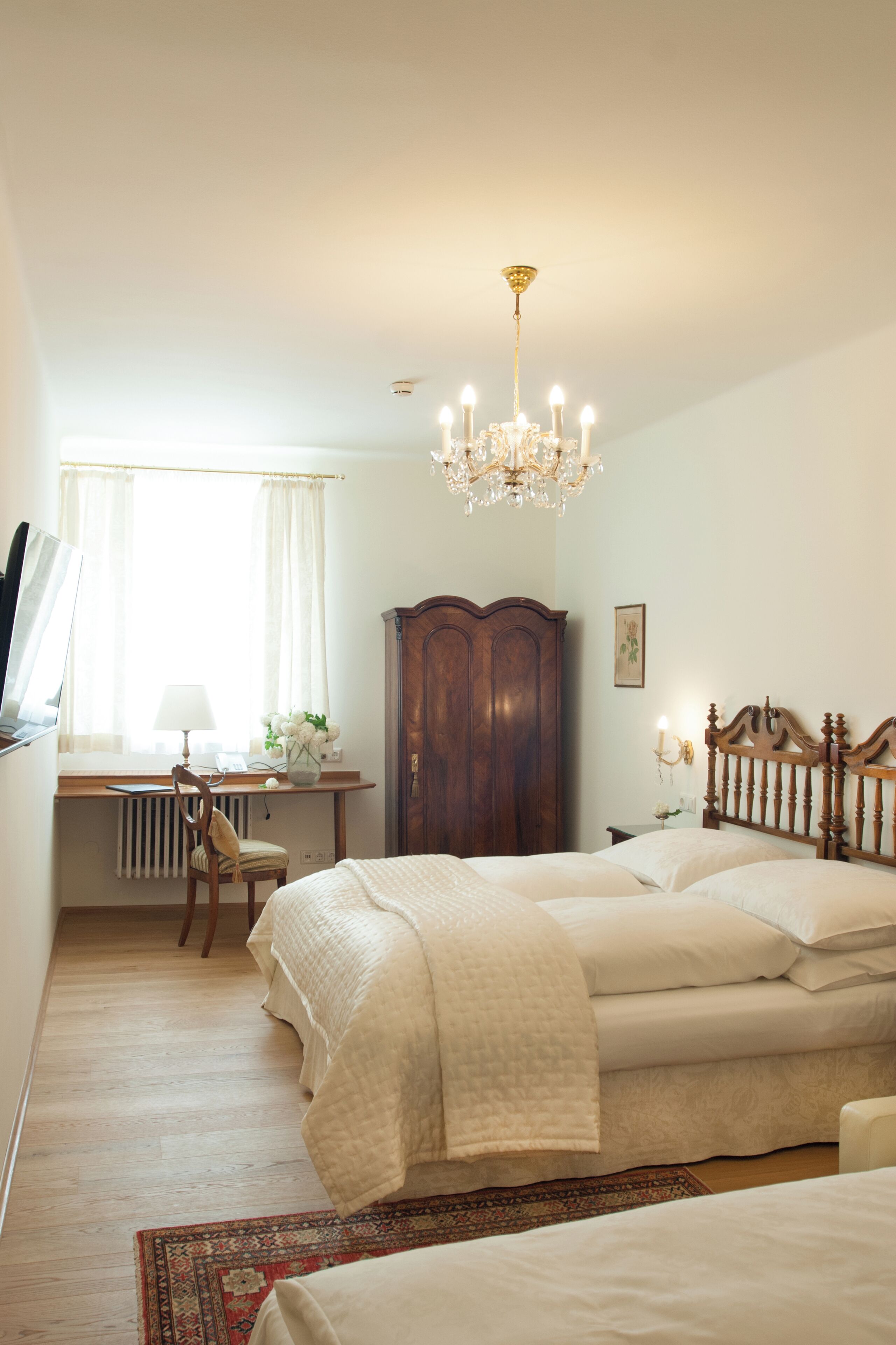 A bedroom with a large bed, a wooden wardrobe, a desk with a chair, and a chandelier.
