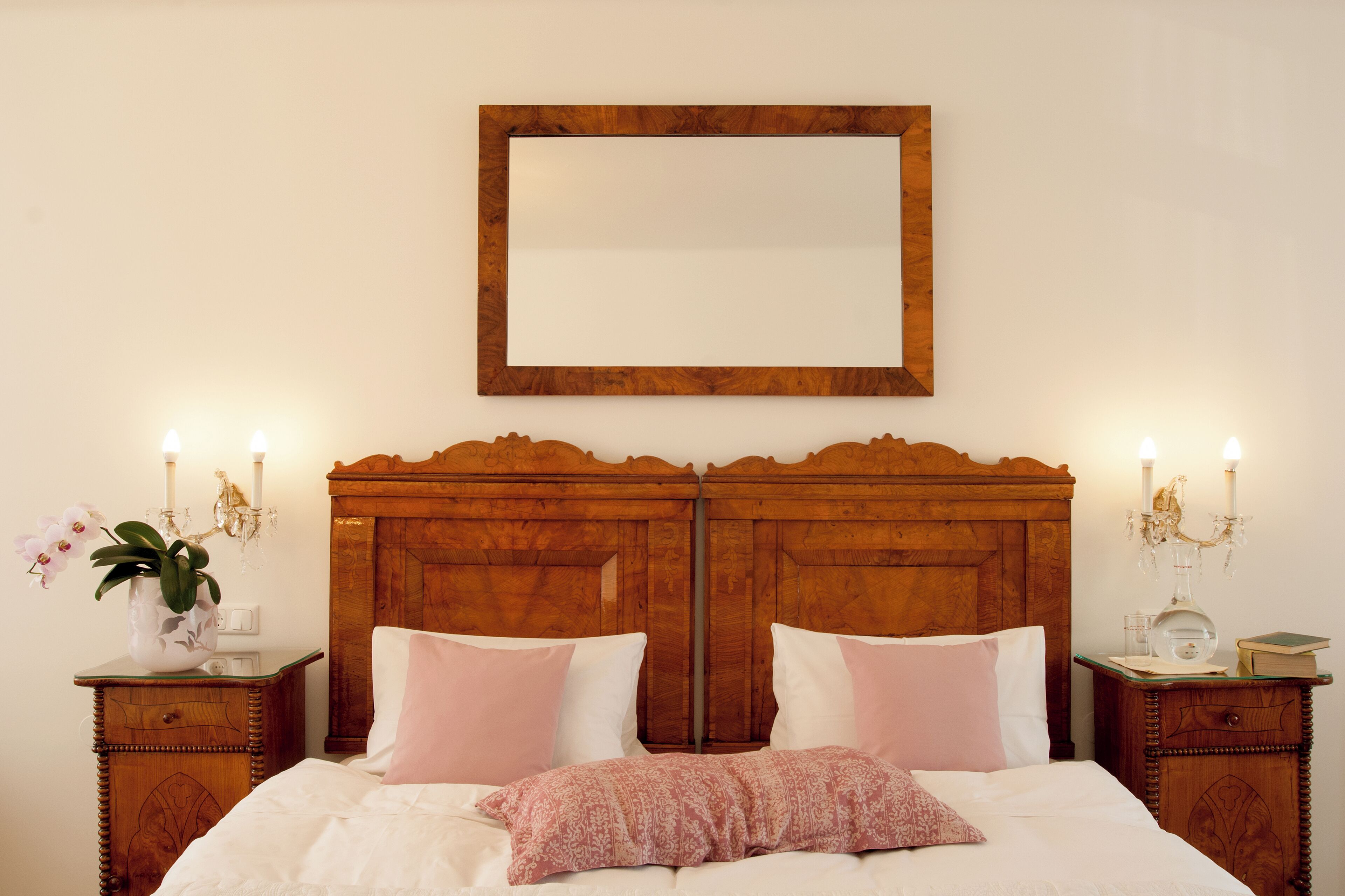 Una cama bien hecha con sábanas blancas y almohadas rosas, con mesitas de noche de madera que sostienen un florero y un libro.