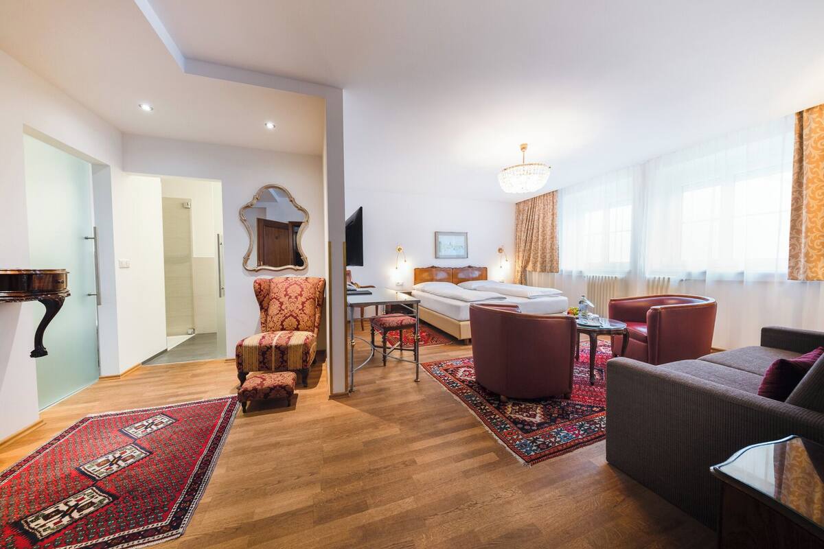 A modern hotel room with a wooden floor, a large bed, a desk, a chair, a sofa, and a TV.