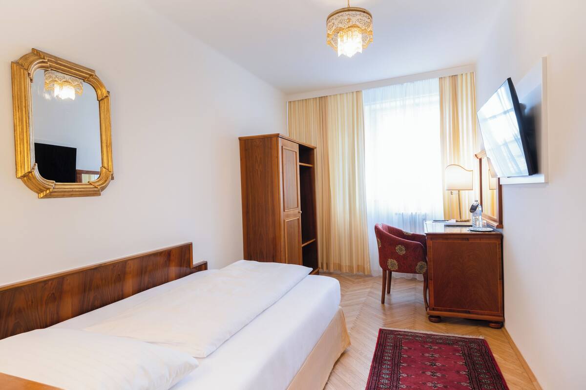 A hotel room with a bed, a wardrobe, a desk with a television, a mirror, and a chandelier.