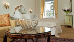A hotel room with a glass coffee table, a bed with a white bedspread, a sofa with a patterned throw, a decorative mirror, and a view of buildings through a window with curtains.